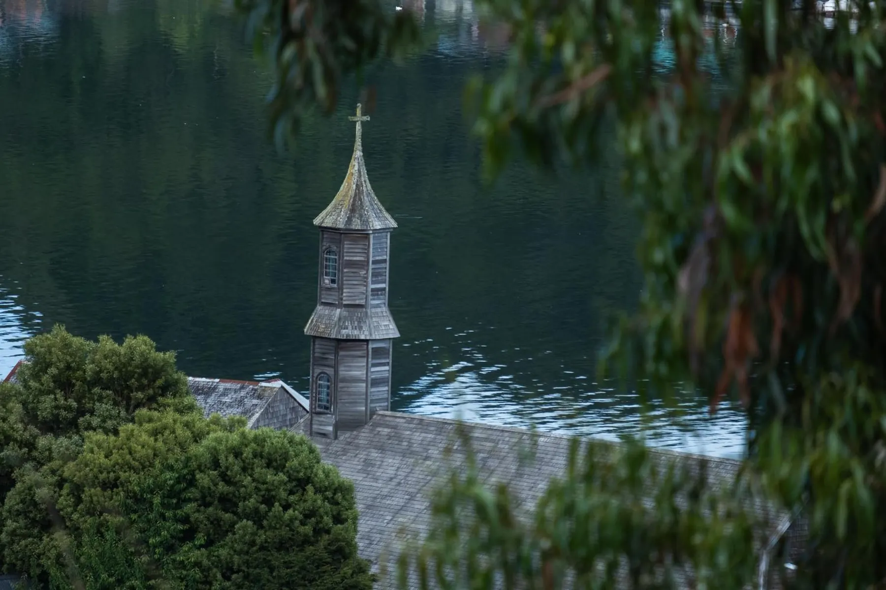 Torre de una iglesia chilota vista entre las ramas, frente al agua